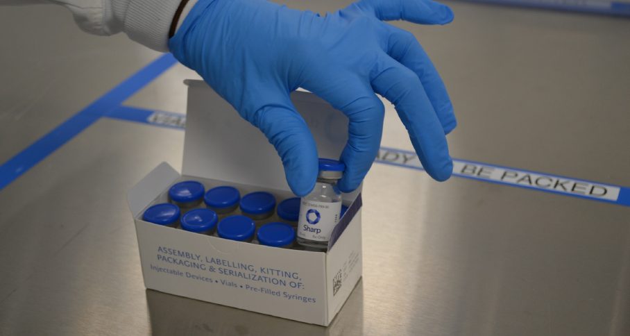 A gloved hand placing a labelled vial into a carton of blue-capped vials ready for packing