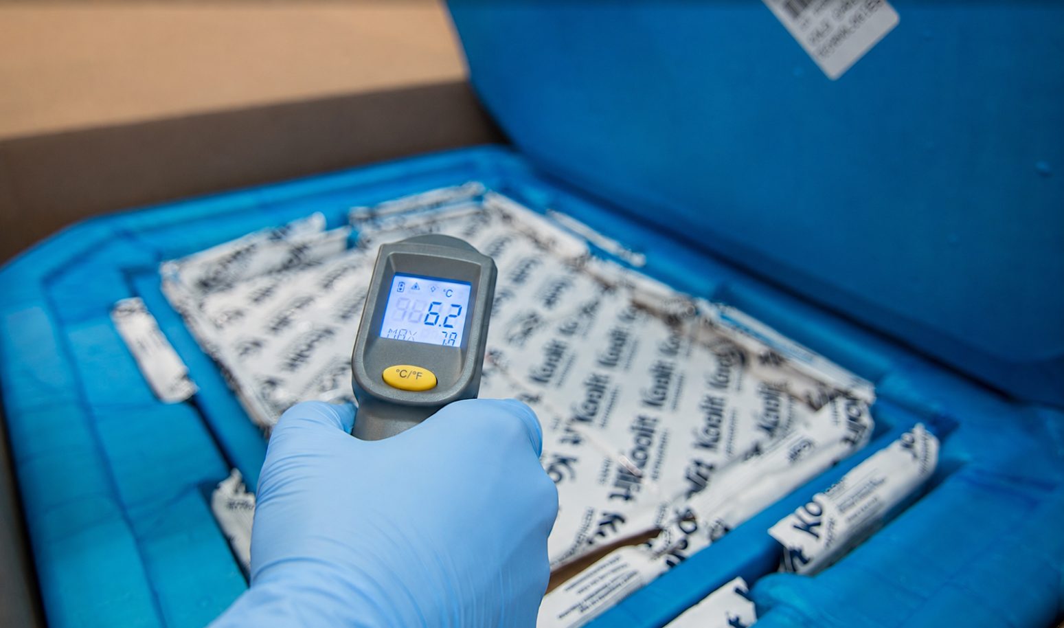 A gloved hand checking an insulated shipper with an infrared thermometer reading 6.2 degrees Celsius