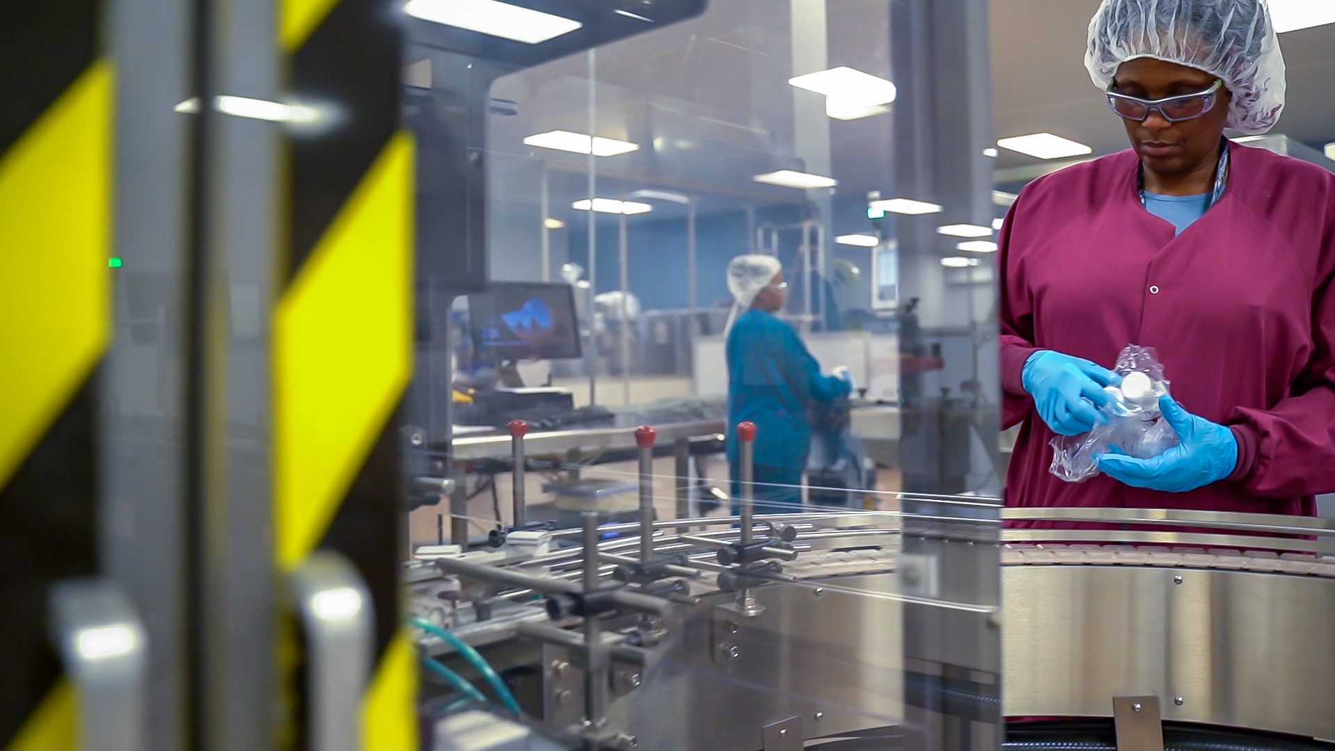 An operator inspecting a bagged bottle beside a packaging conveyor, with a colleague working behind